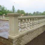 Natural stone balustrade with pillars on a raised platform, surrounded by grass and trees under a cloudy sky.