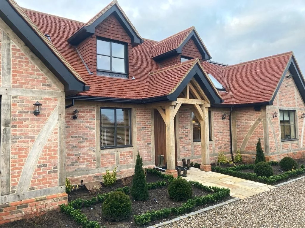 A modern brick house with a red tiled roof, wooden beams, dormer windows, and landscaped front garden with small shrubs and gravel path.
