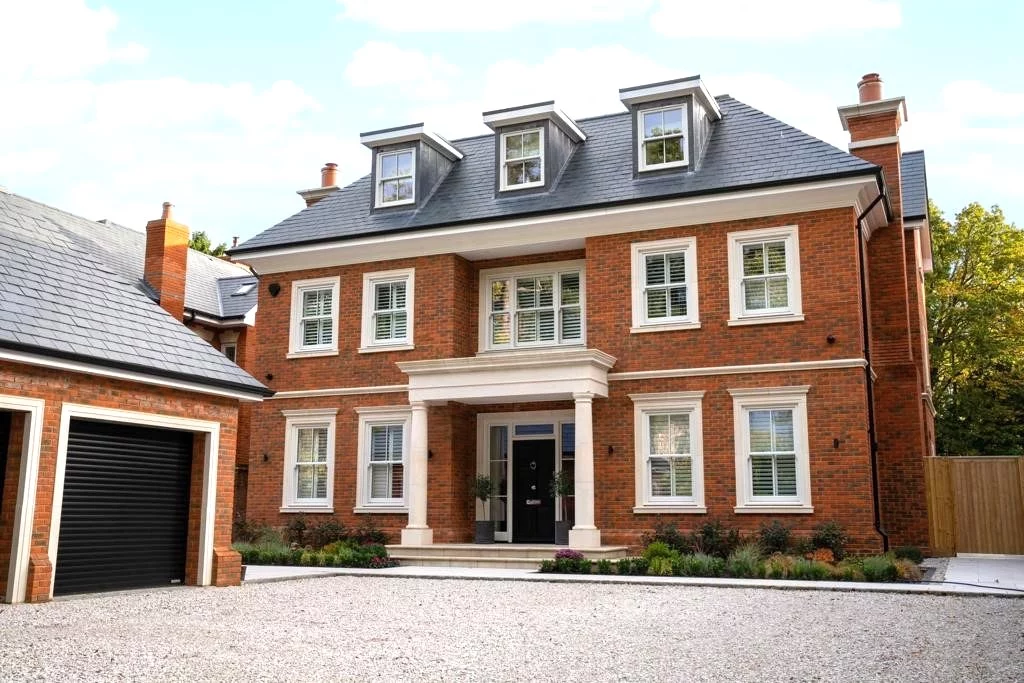 A large, three-story red brick house with white trim, dormer windows, a columned entrance, and a gravel driveway. A detached garage is on the left.