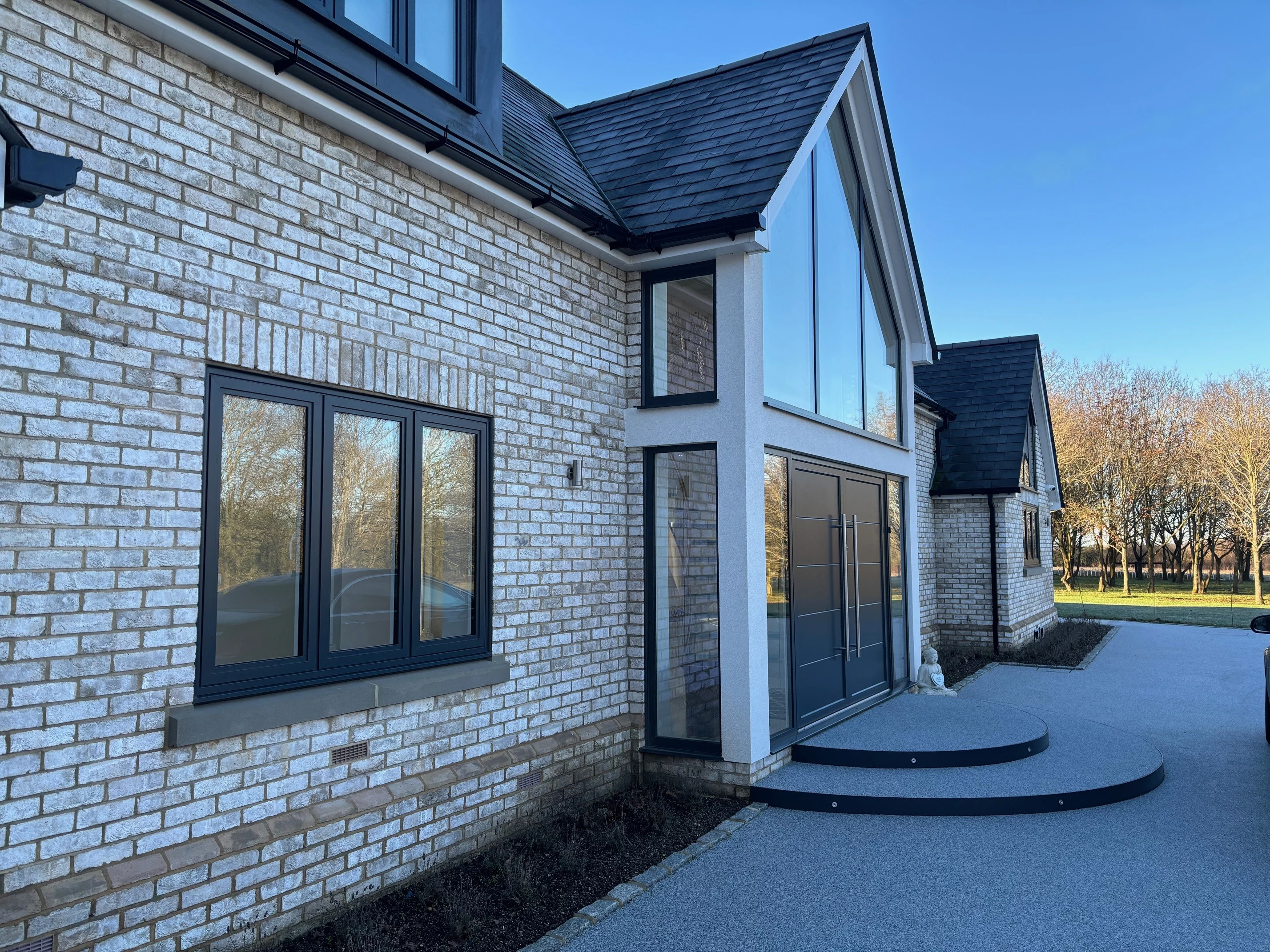 Modern two-story house with white brick exterior, large tinted windows, double front doors, and curved steps, set against a clear blue sky and a driveway.