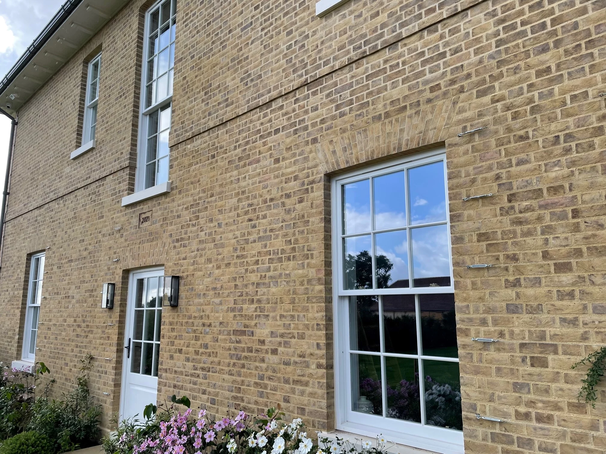 A brick house with white-framed windows and door, a wall-mounted light fixture, and a flower bed in front. Blue sky and greenery are reflected in the windows.