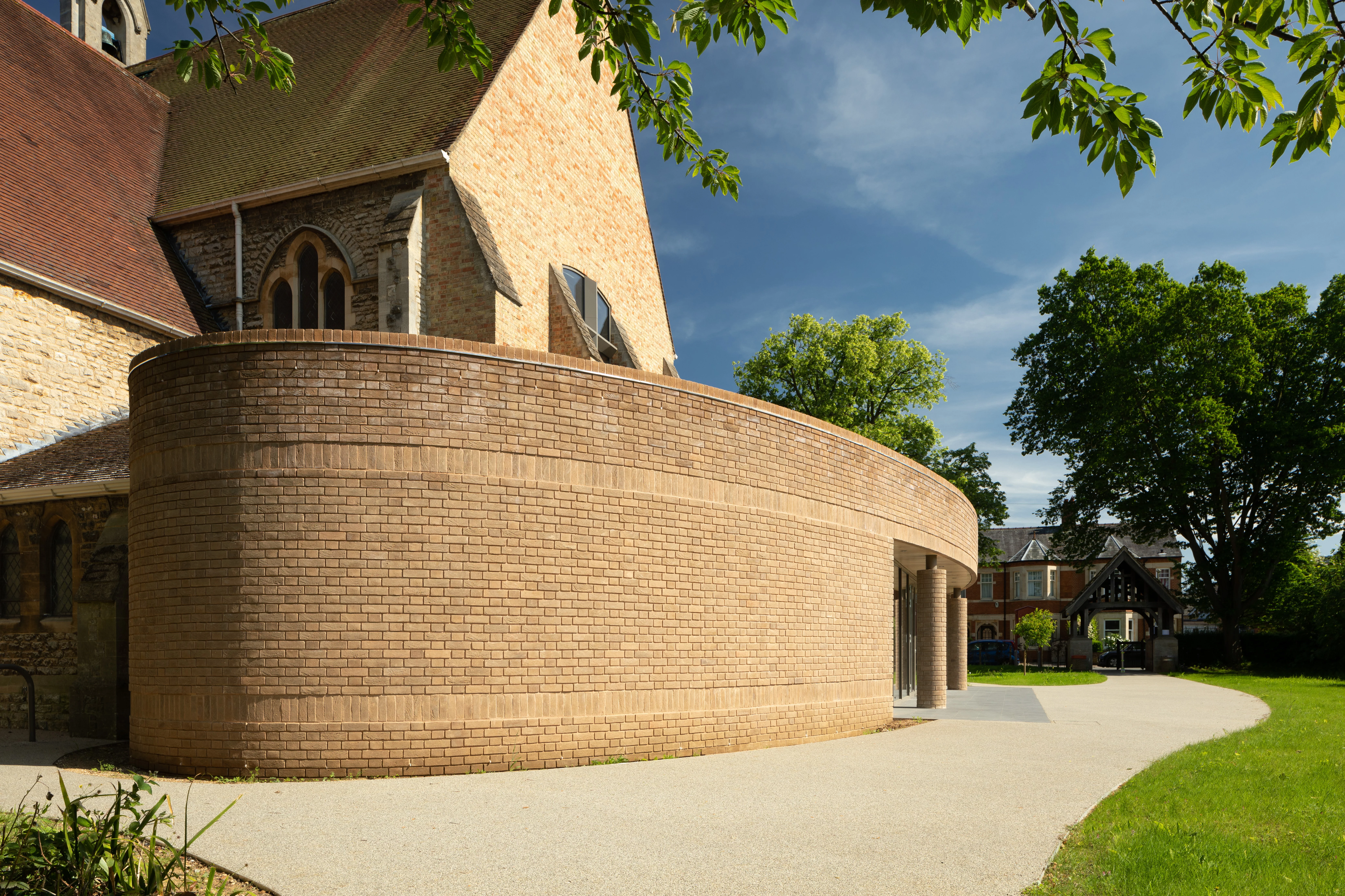 Curved brick extension attached to a historic stone church, with a paved path and trees under a blue sky.