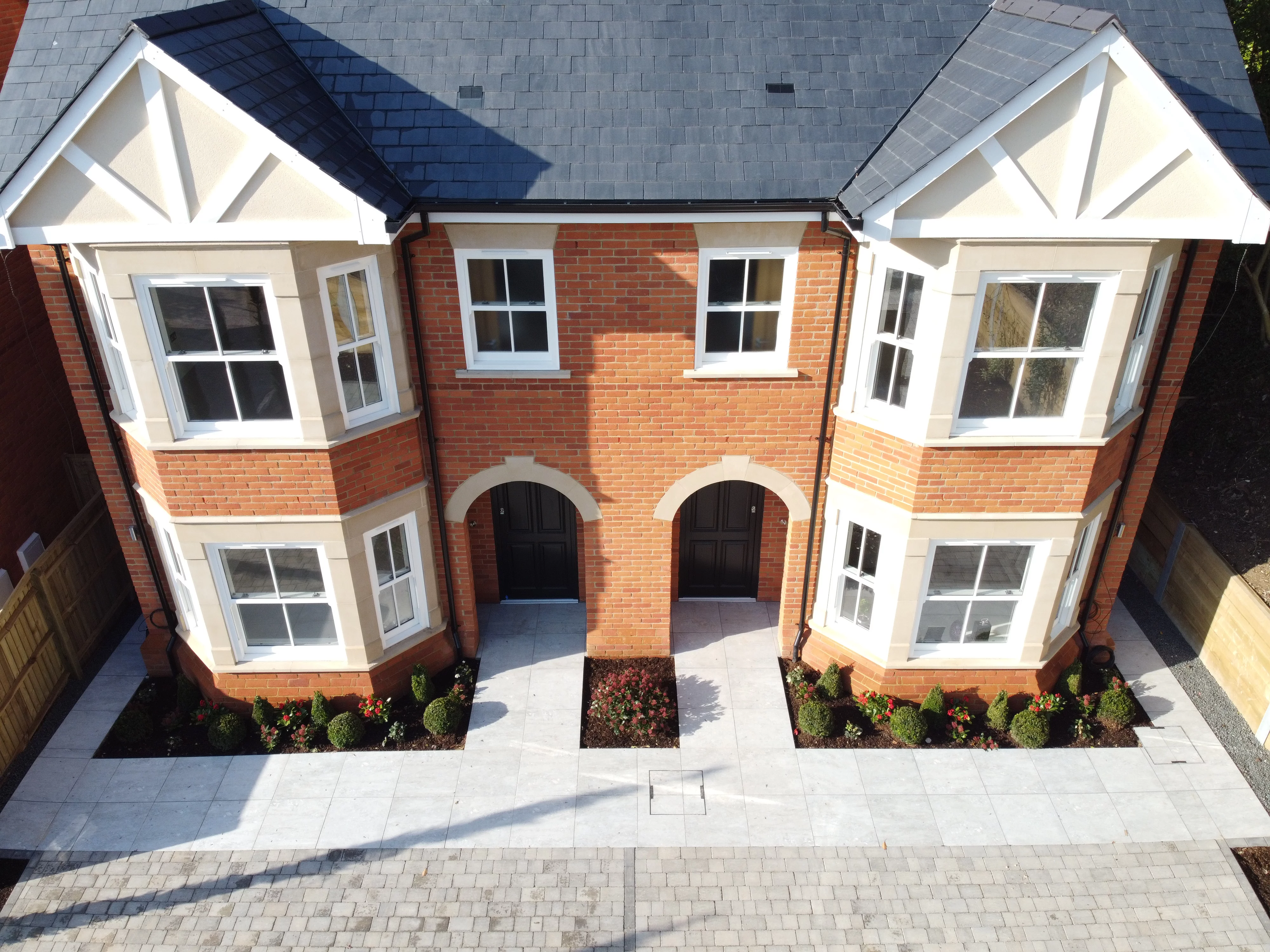 Aerial view of a modern semi-detached brick house with two front doors, large bay windows, and a paved walkway surrounded by small shrubs.