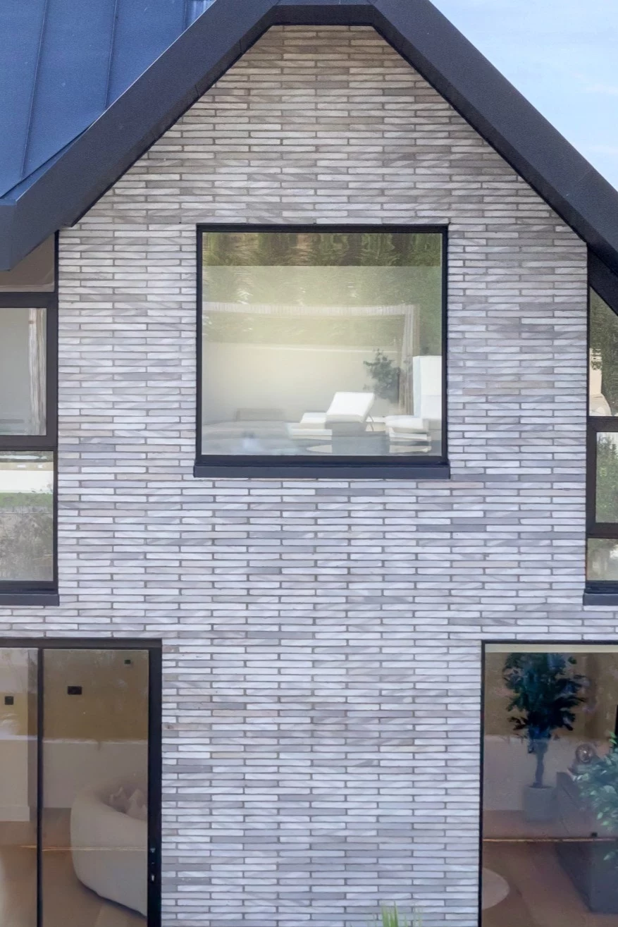 Modern house exterior with grey linear brick facade, large square window on upper floor, and floor-to-ceiling windows on the ground floor. Minimalist interior furniture visible through windows.