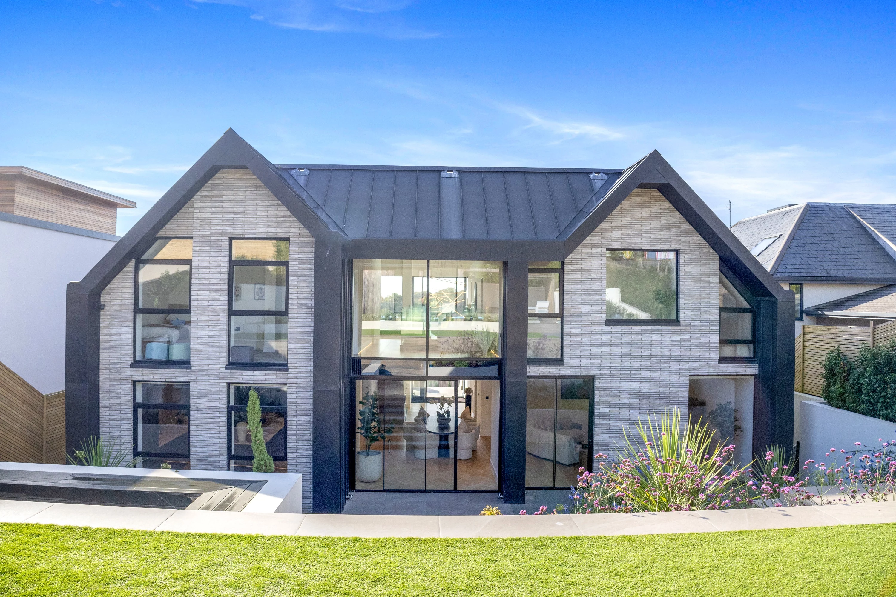 Modern two-story house with large windows, light linear brick exterior, black roof, and landscaped yard under a clear blue sky.