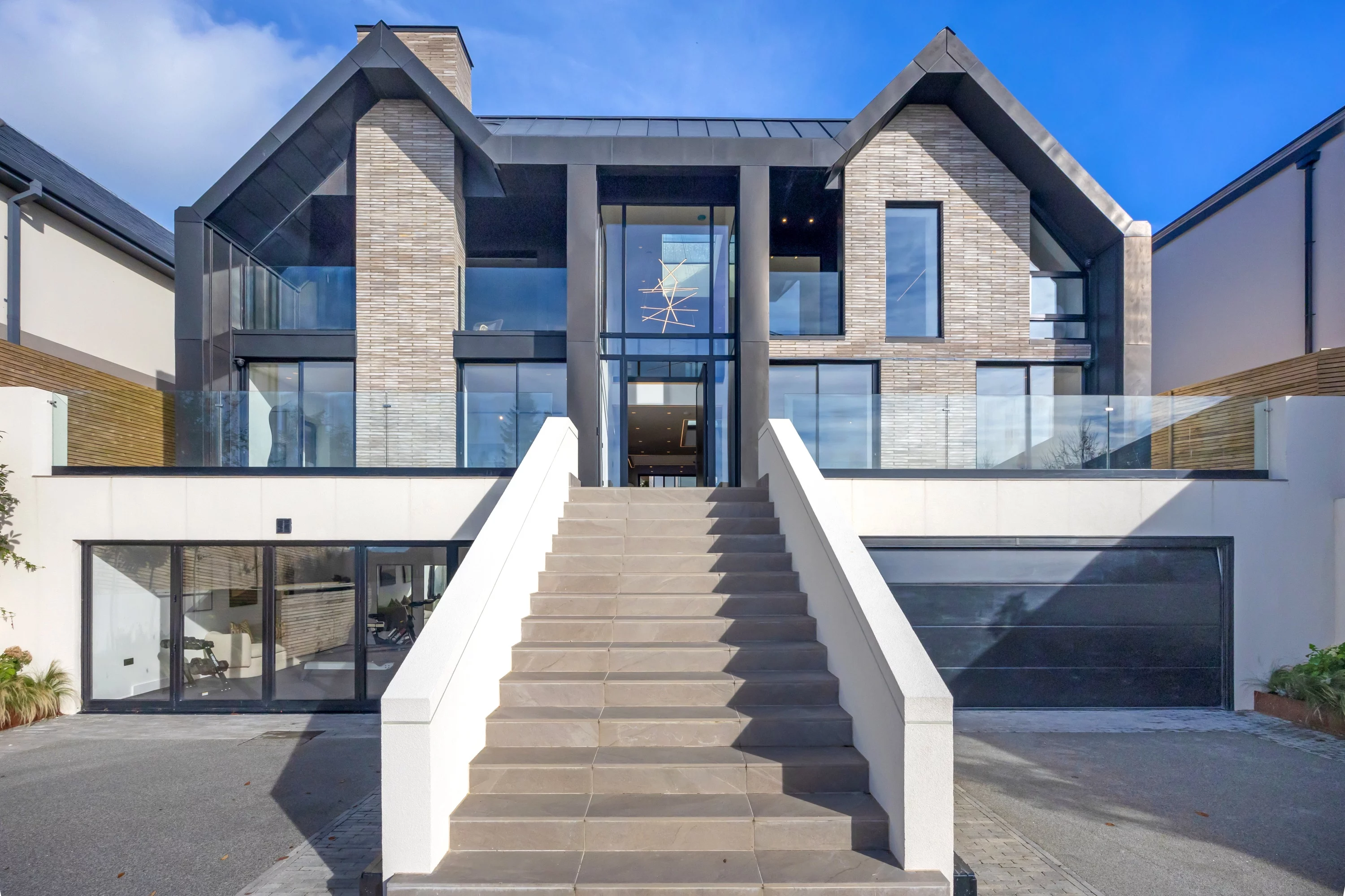 A modern two-story house with large windows, a linear brick facade, wide front steps, and a double garage under a blue sky.