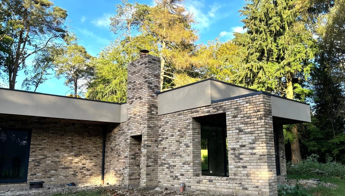 A modern single-story house with beige and gray brick exterior, large windows, and a chimney, surrounded by tall green trees under a blue sky.