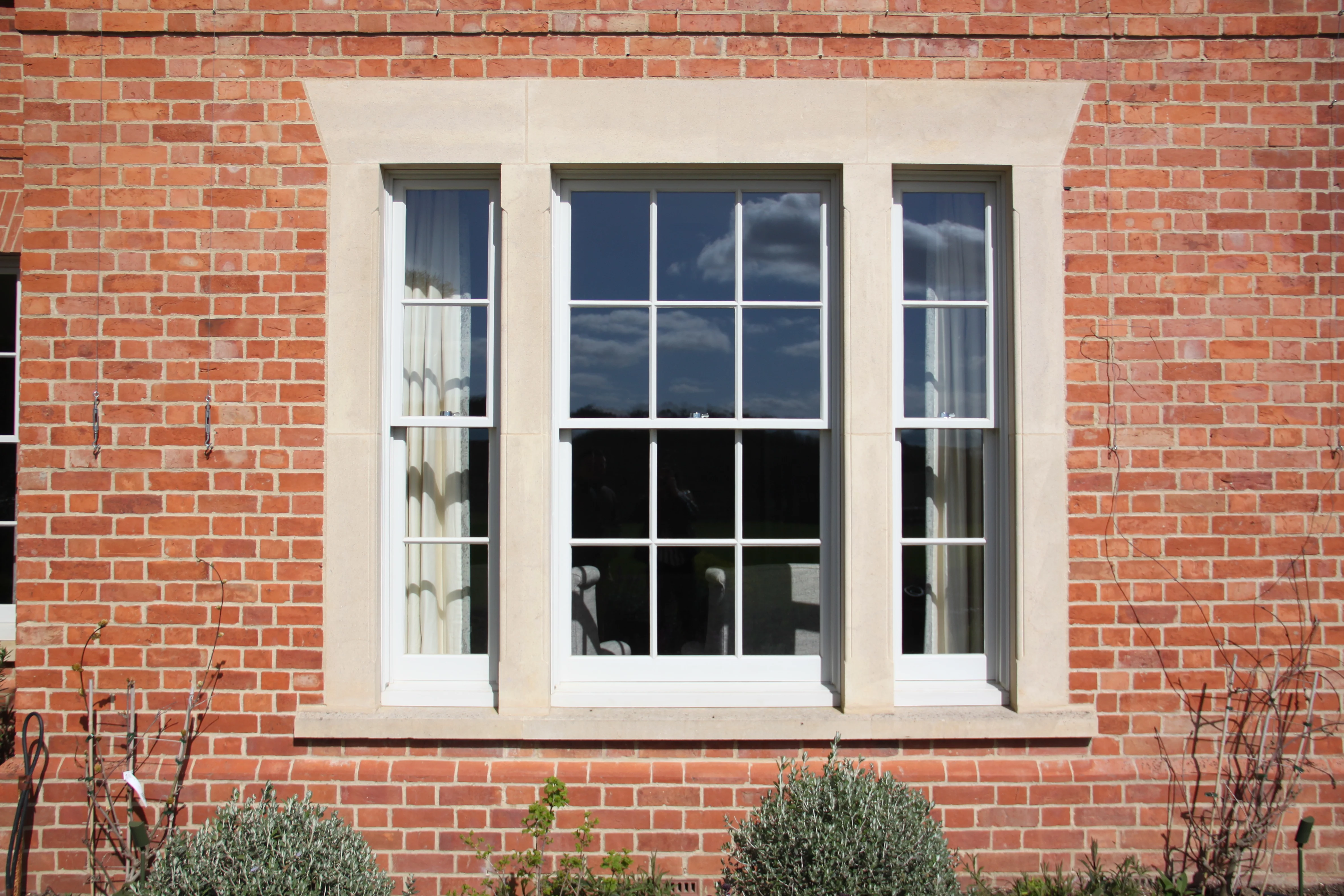 A large rectangular window with white frames set in a red brick wall, reflecting the sky and clouds, with plants at the base.