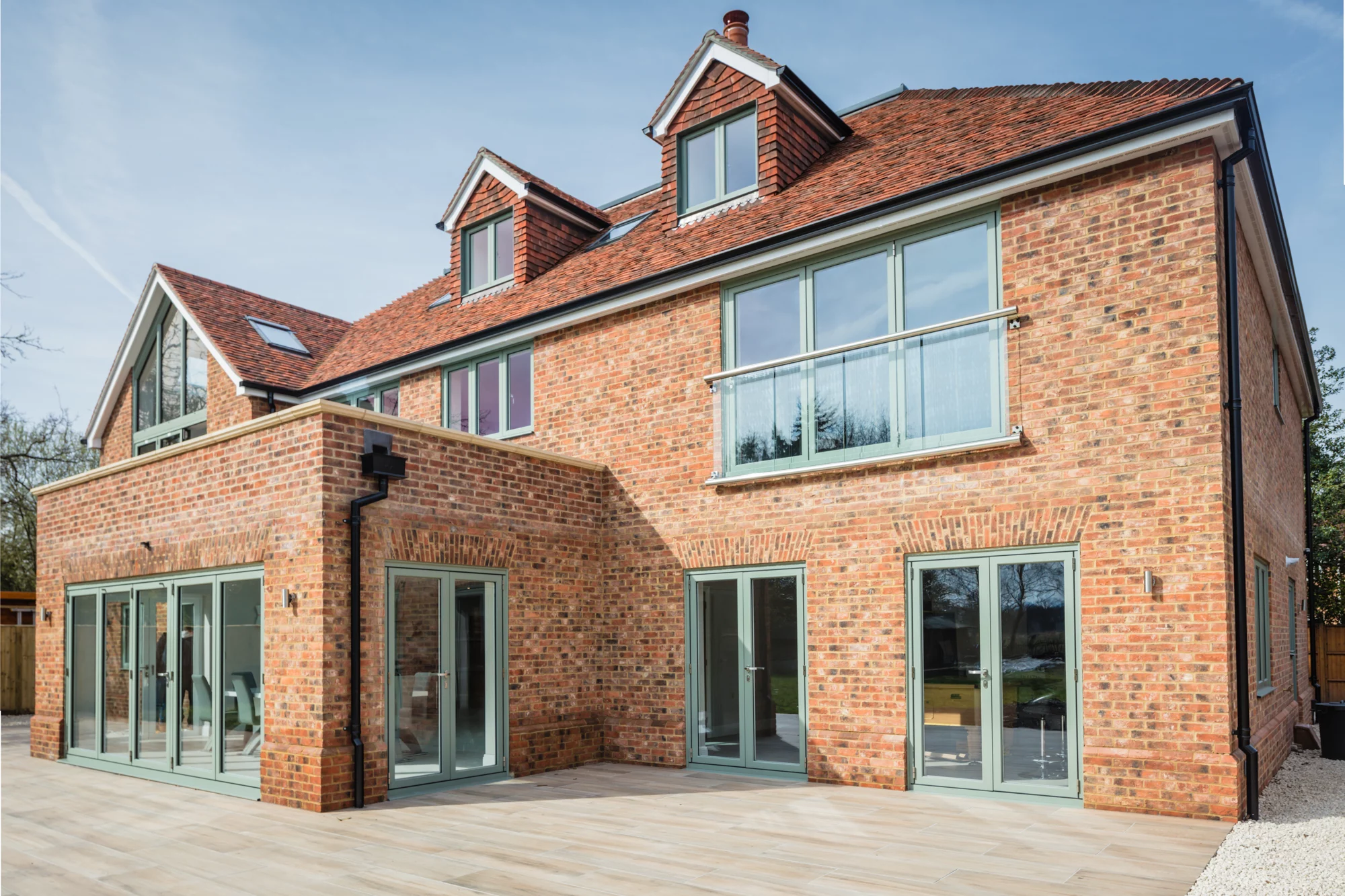 A modern red-brick house with large windows, glass doors, and a sloped tiled roof, featuring two upper dormer windows and a spacious patio area.