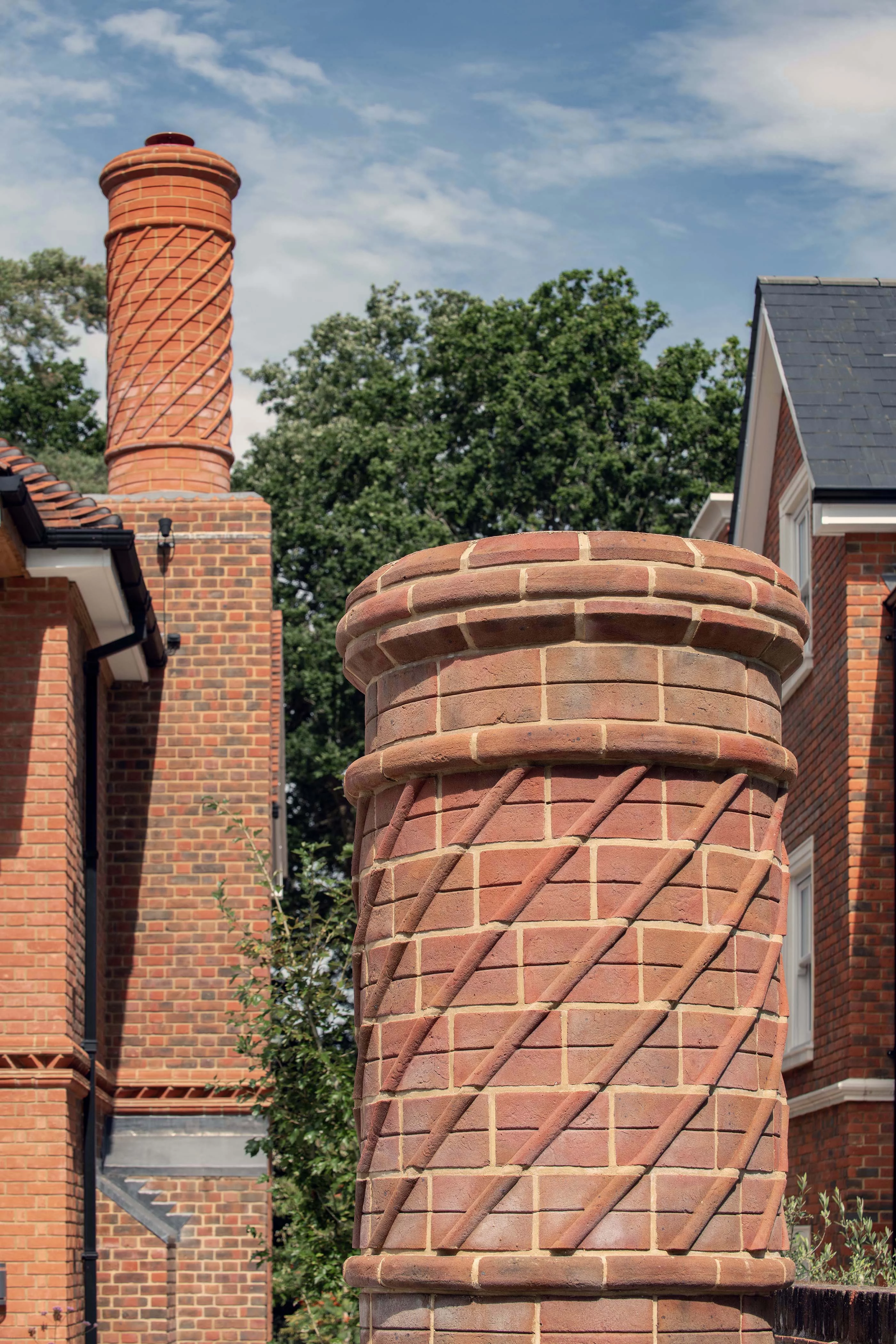 A close-up of a decorative brick chimney with a spiral pattern on a house exterior, with a similarly patterned chimney and trees in the background.