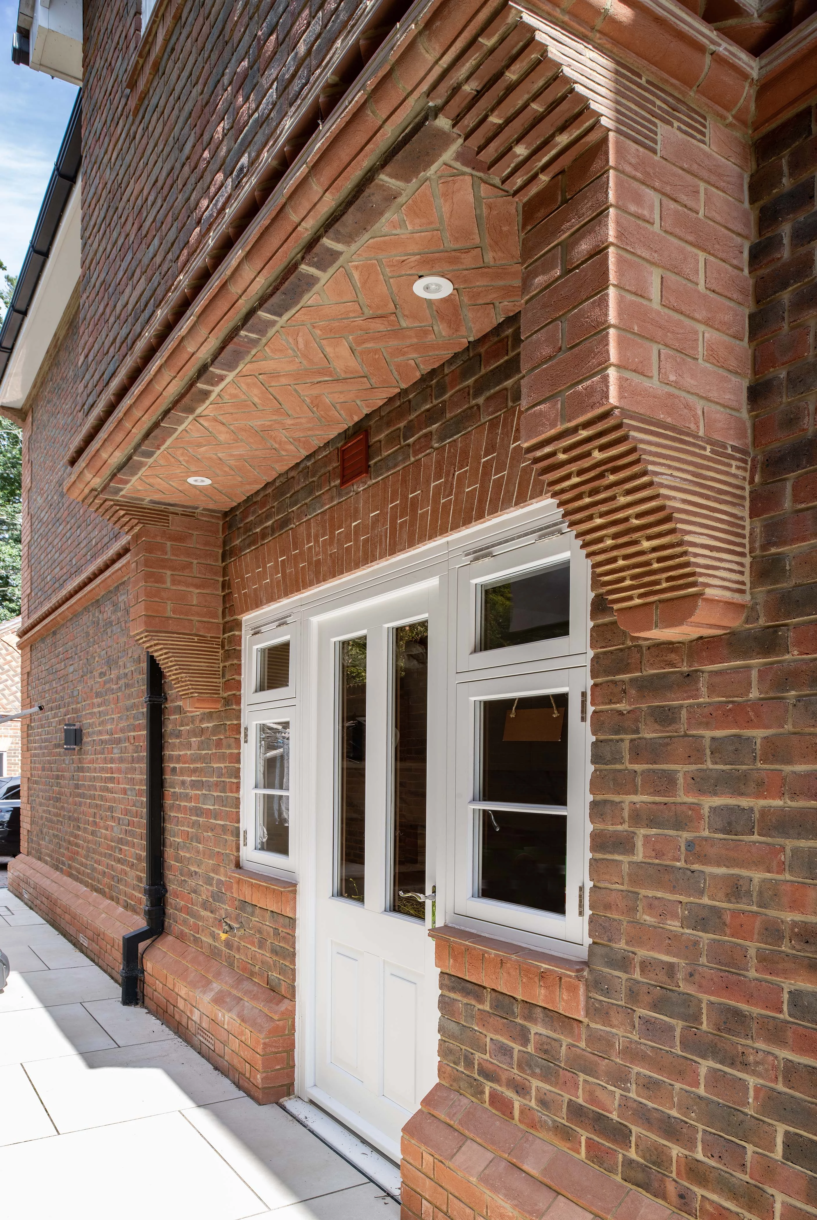A close-up view of a building facade with intricate brickwork around a white door and two windows, featuring a covered entryway and paved walkway.
