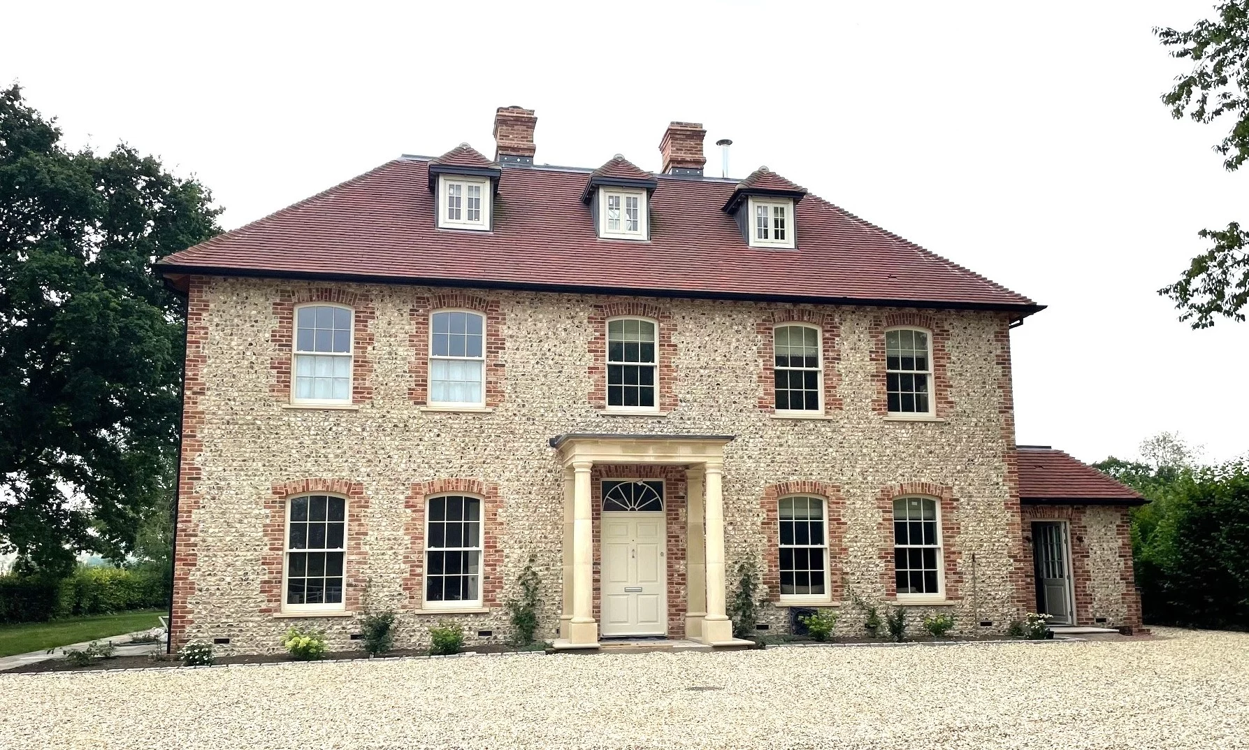 Two-storey loose flint faced house with red roof, symmetrical windows, central front door with columns, and gravel drive in front.