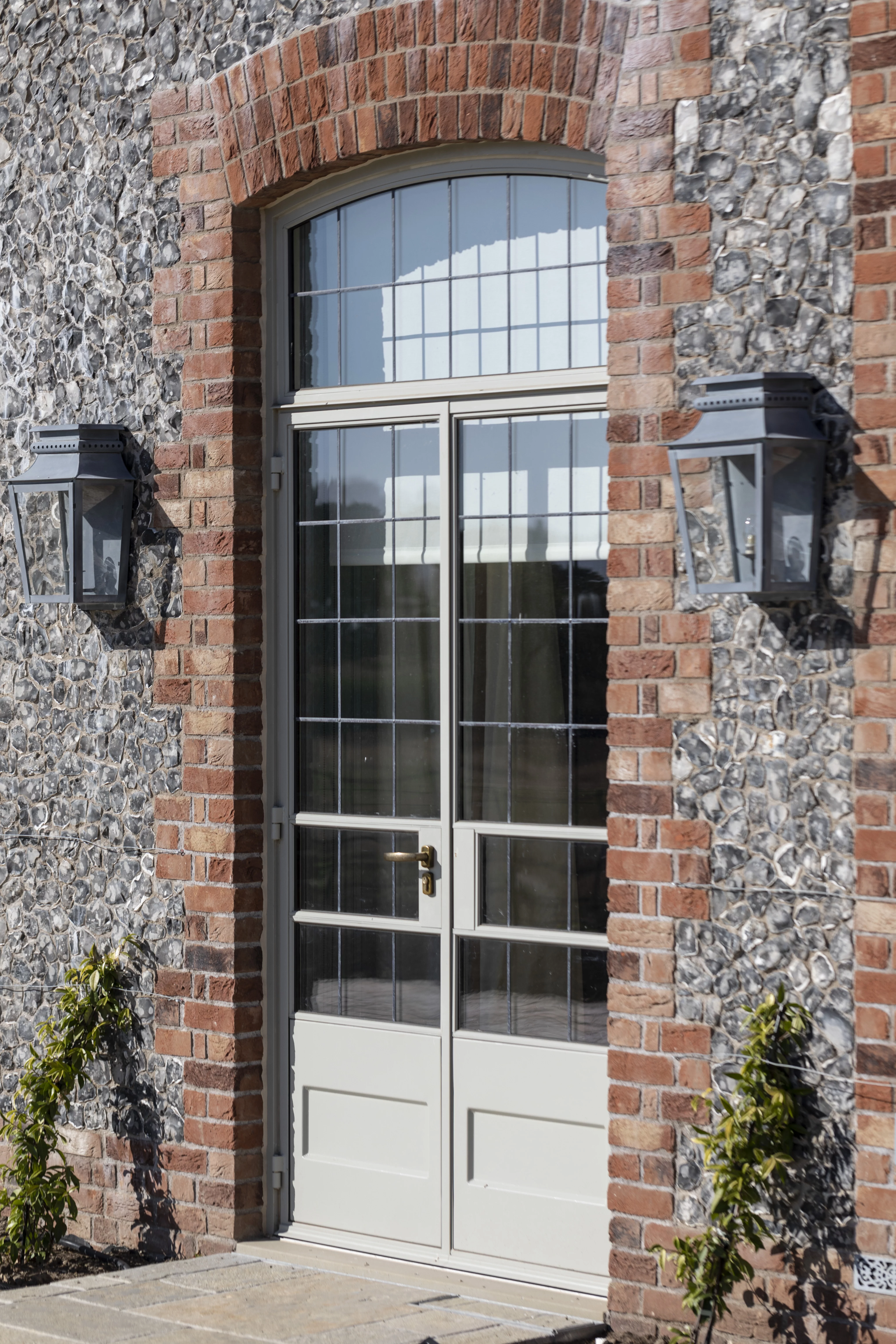 A glass-paned double door with a metal handle is set in a brick and stone wall, flanked by two black wall lanterns and small green shrubs.