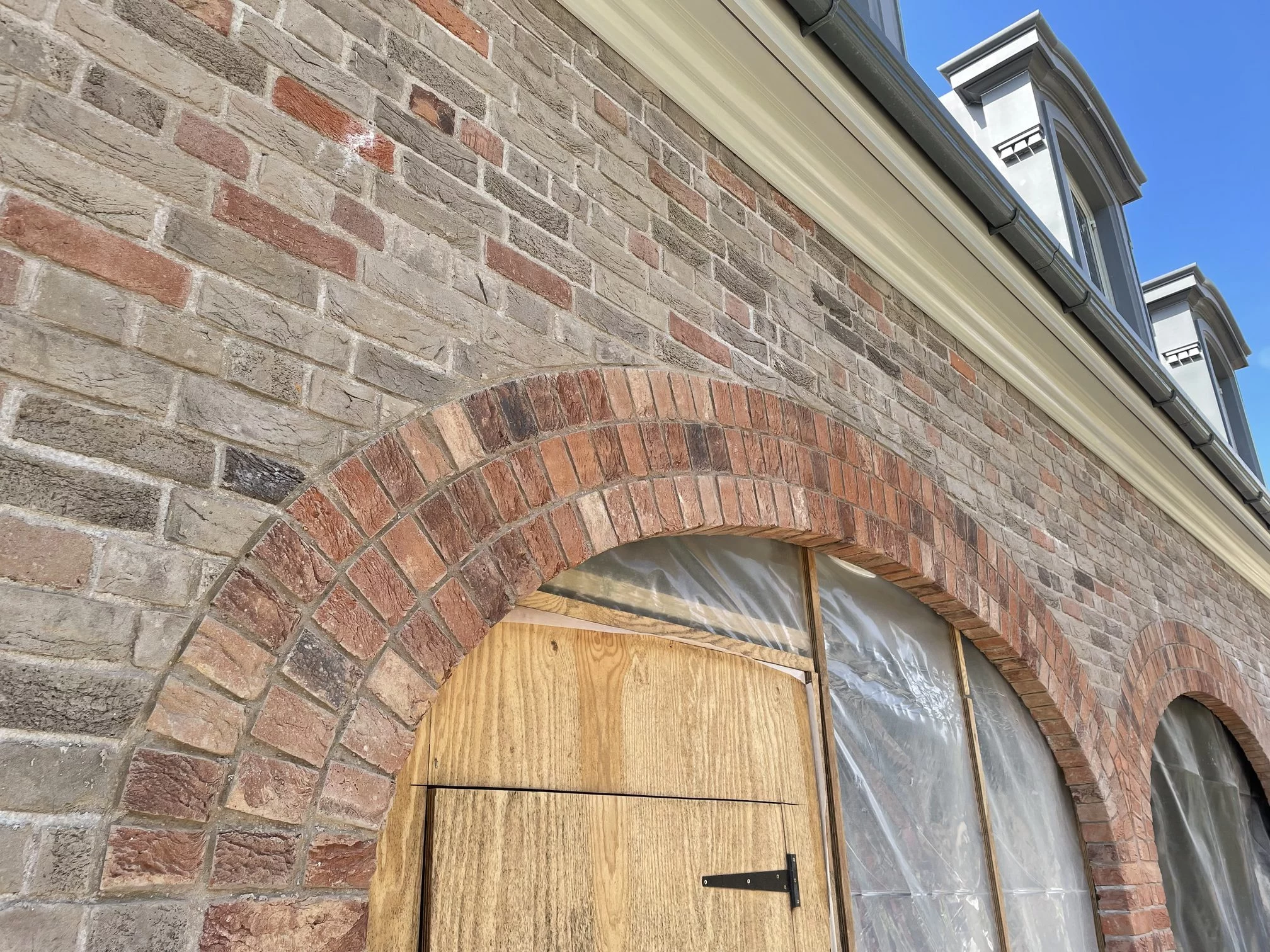 Brick building façade with arched brickwork above a wooden door and plastic-covered windows; upper storey features dormer windows with grey trim.