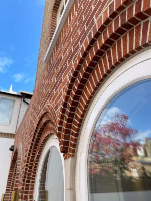 An angled shot of a pair of windows both curved at the top with elliptical pre-fabricated brick arches featuring at the top with reflections of pink trees int eh window and blue skies