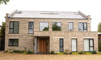 Grey linear brick house Modern two-storey house with linear light brick exterior, large rectangular windows, and a wooden front door, set on a gravel drive with small landscaped plants.