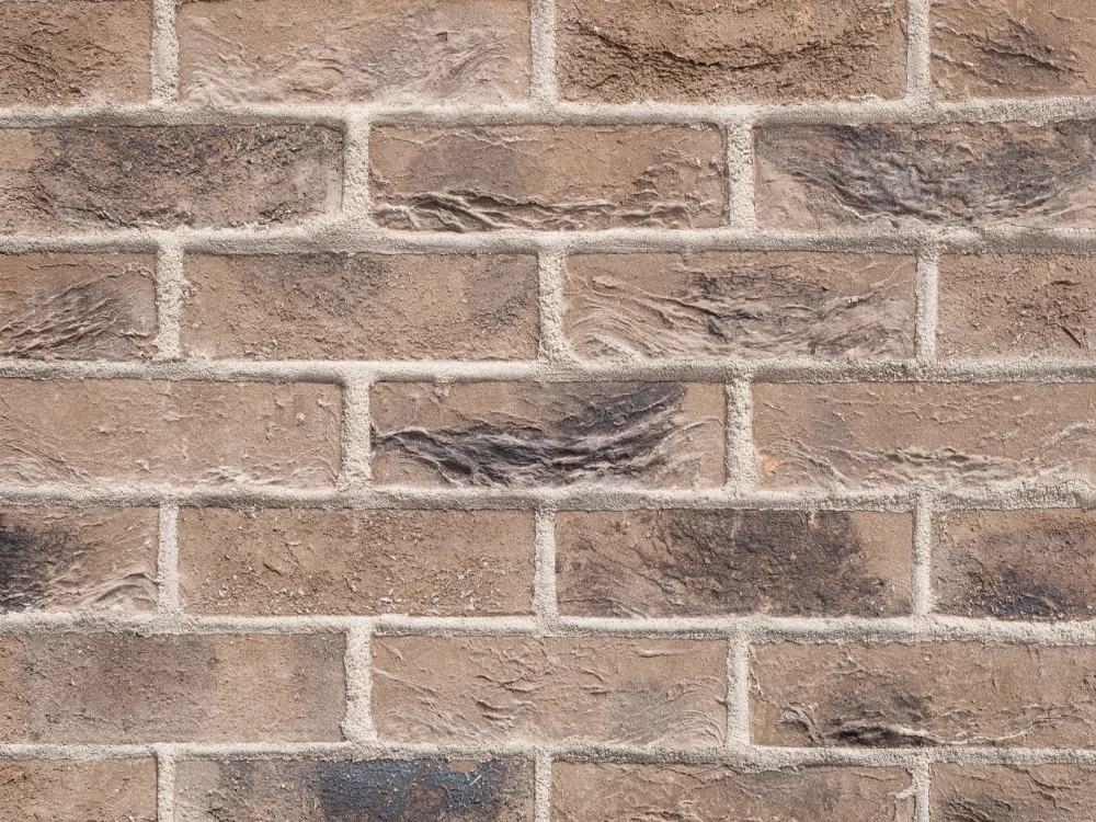 Merino Grey, a brown brick wall featuring horizontal and vertical mortar lines; the bricks show a rough, textured surface with varied colour tones.