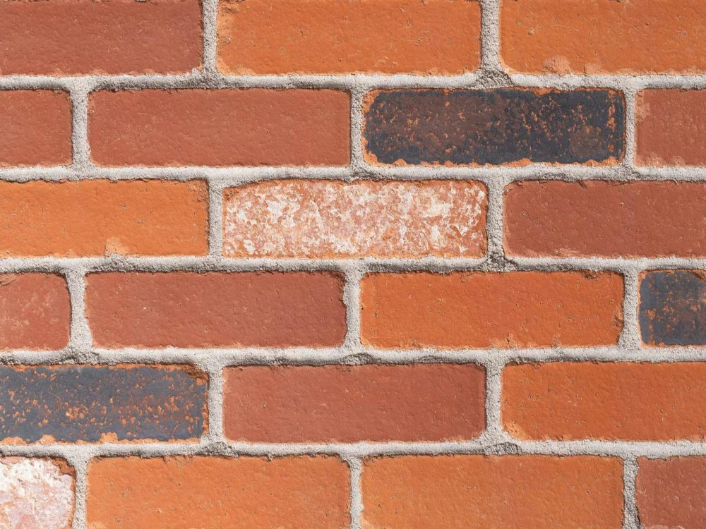 A red brick wall with grey mortar, highlighting the varied shades and textures of the bricks in a horizontal arrangement.