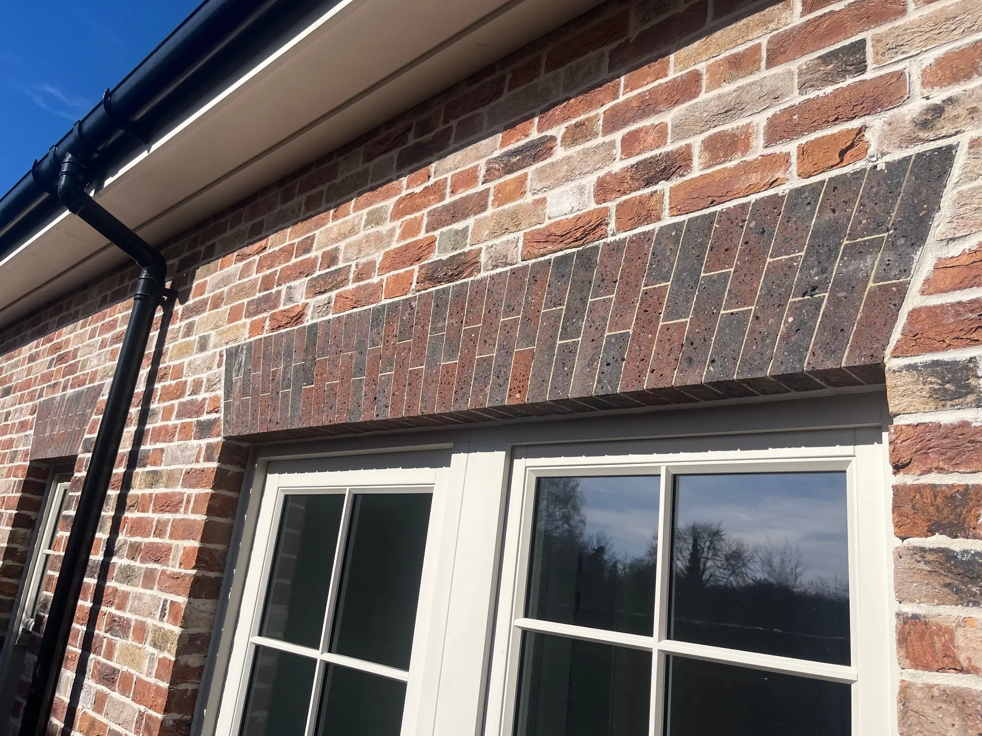 Close-up of a brick house exterior showing two white-framed windows beneath an arch of darker bricks, with a black gutter running along the roofline.