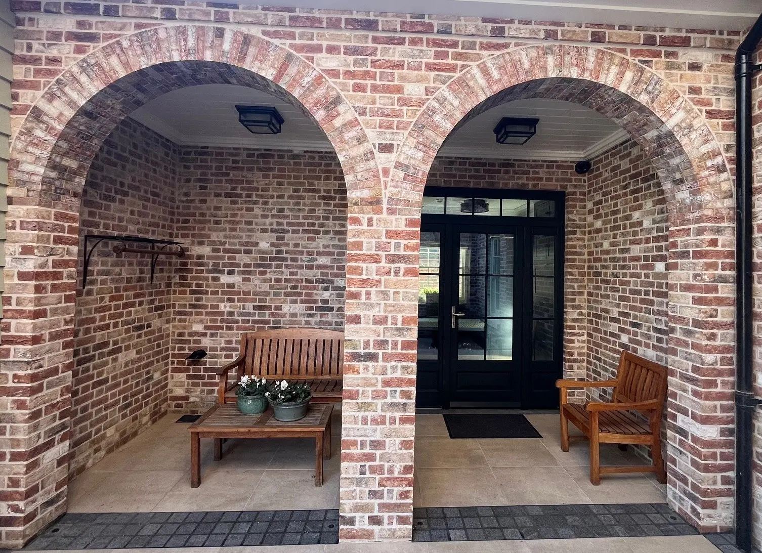 Two wooden benches and potted plants sit under brick archways in a covered entrance with a glass double door.