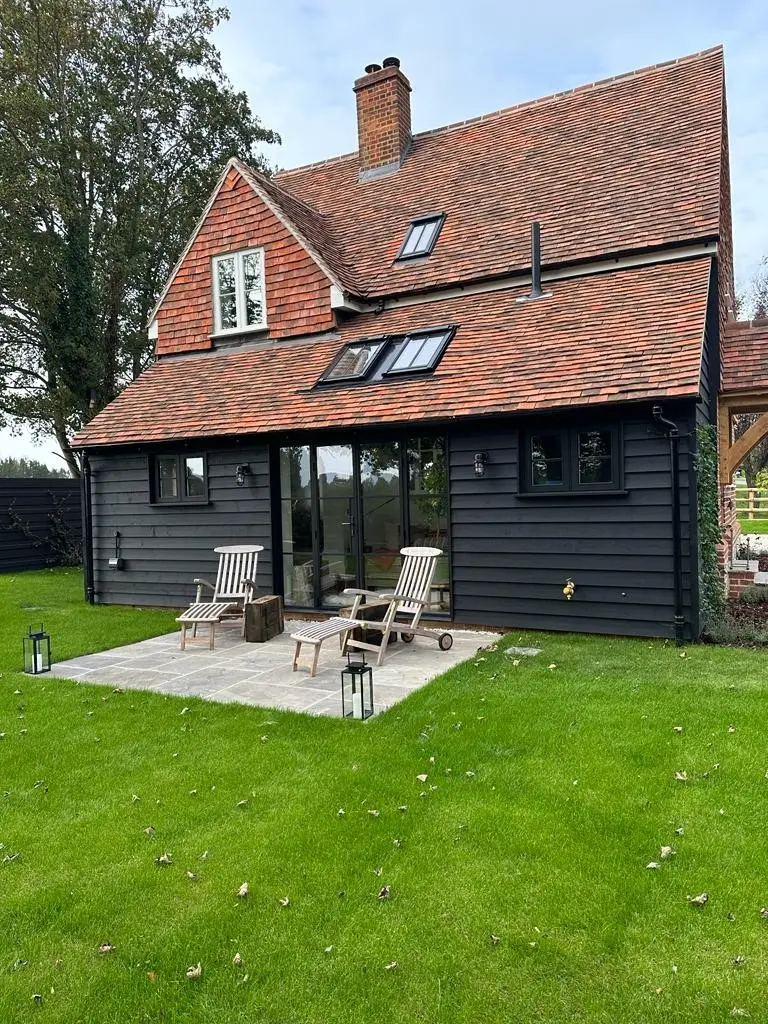 A two-storey house with red roof clay tiles, black wood cladding, large glass doors, a small patio with two lounge chairs, and a lawn with trees in the background.