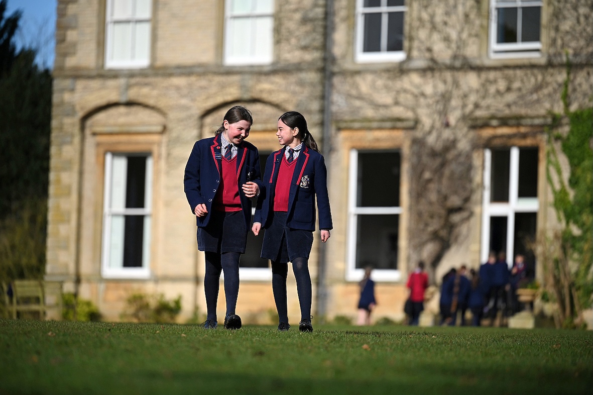 Two girls in school uniform chatting and walking across the lush green lawn at Swanbourne House School, with the picturesque Main House in the background.